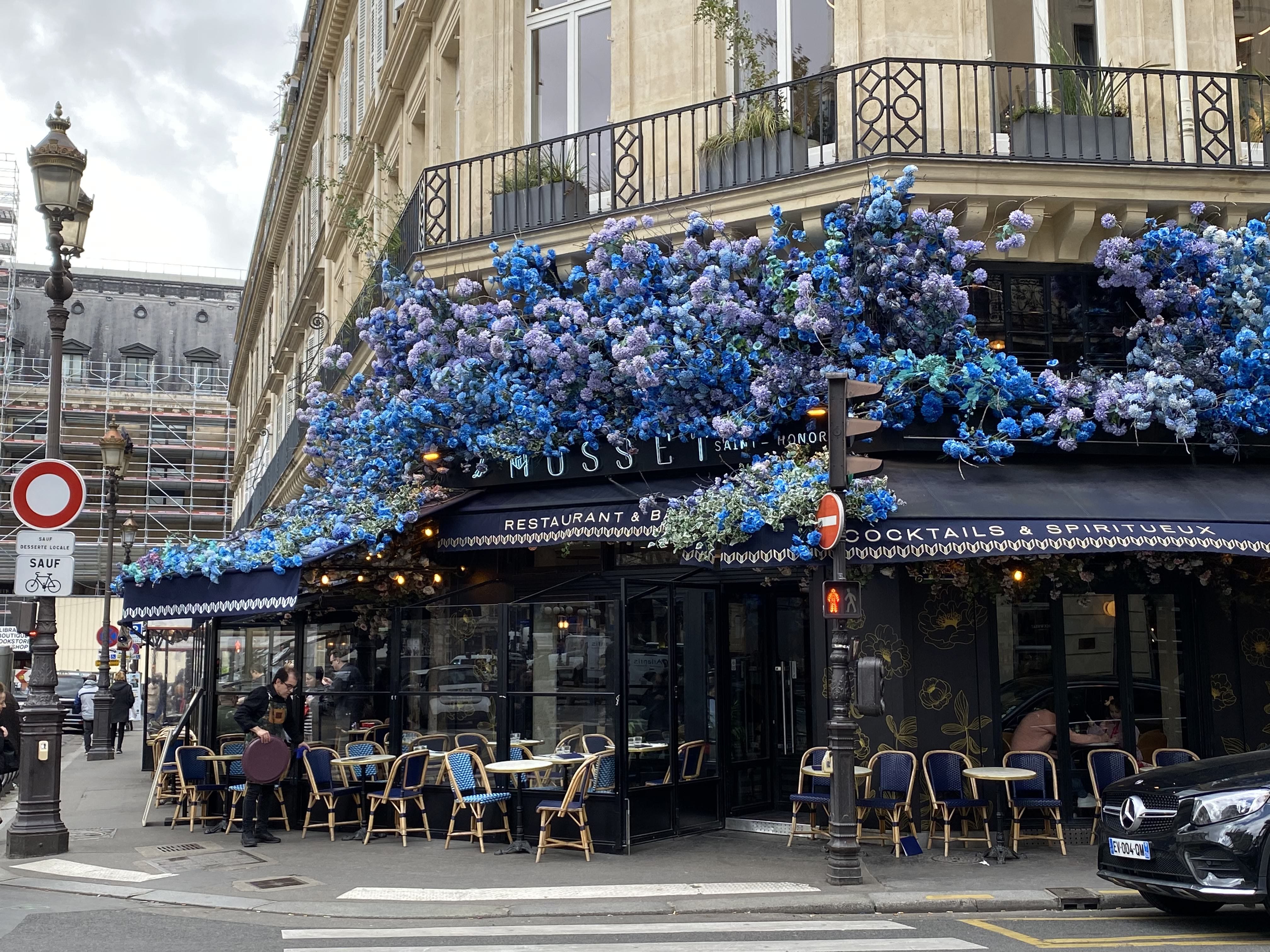 Ein französisches Café an einer Straßenecke mit Sitzgelegenheiten im Freien und blauen Blumen auf der Markise
