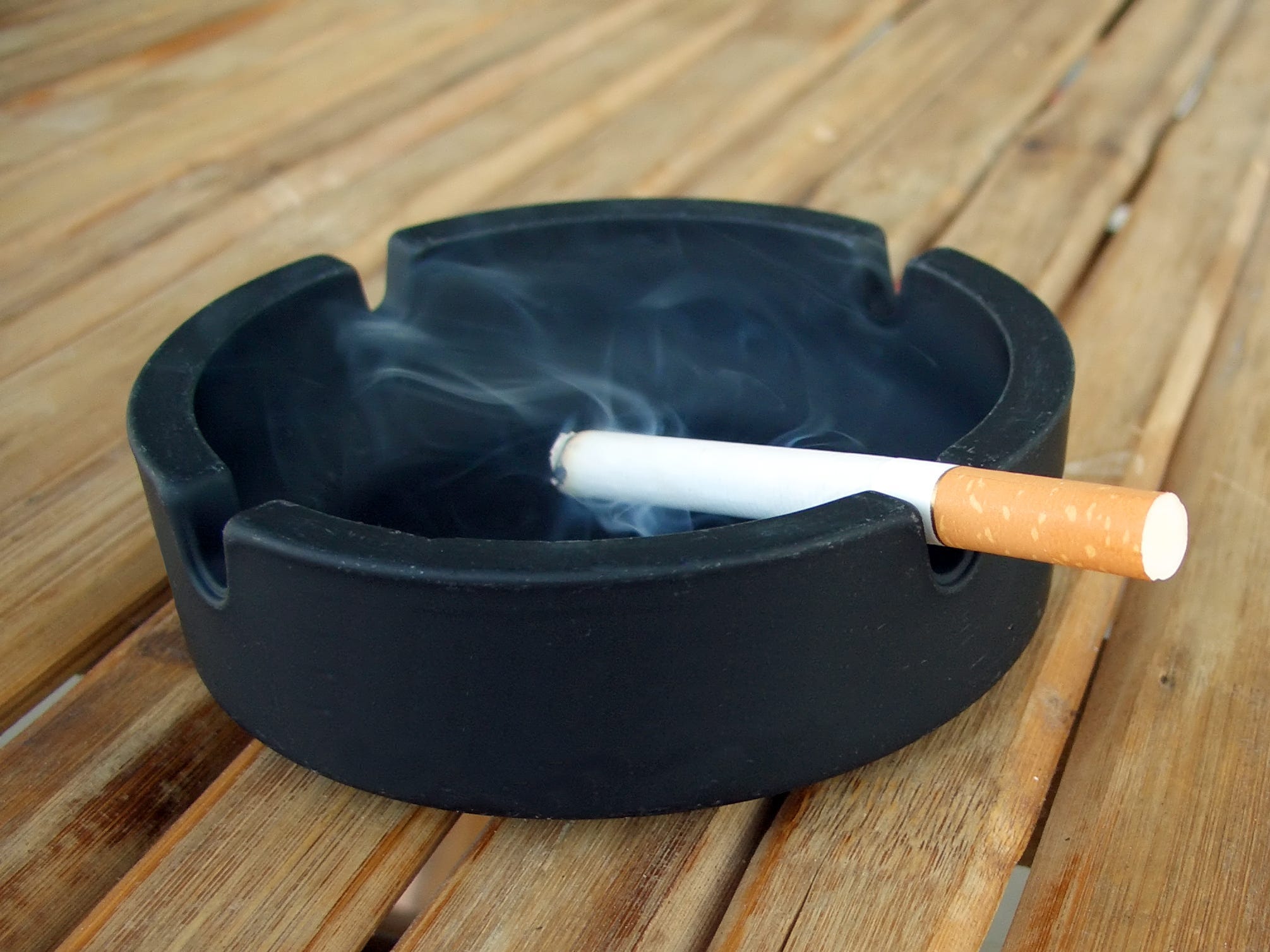 Ein schwarzer Aschenbecher auf einem Holztisch mit einer rauchenden Zigarette darauf.