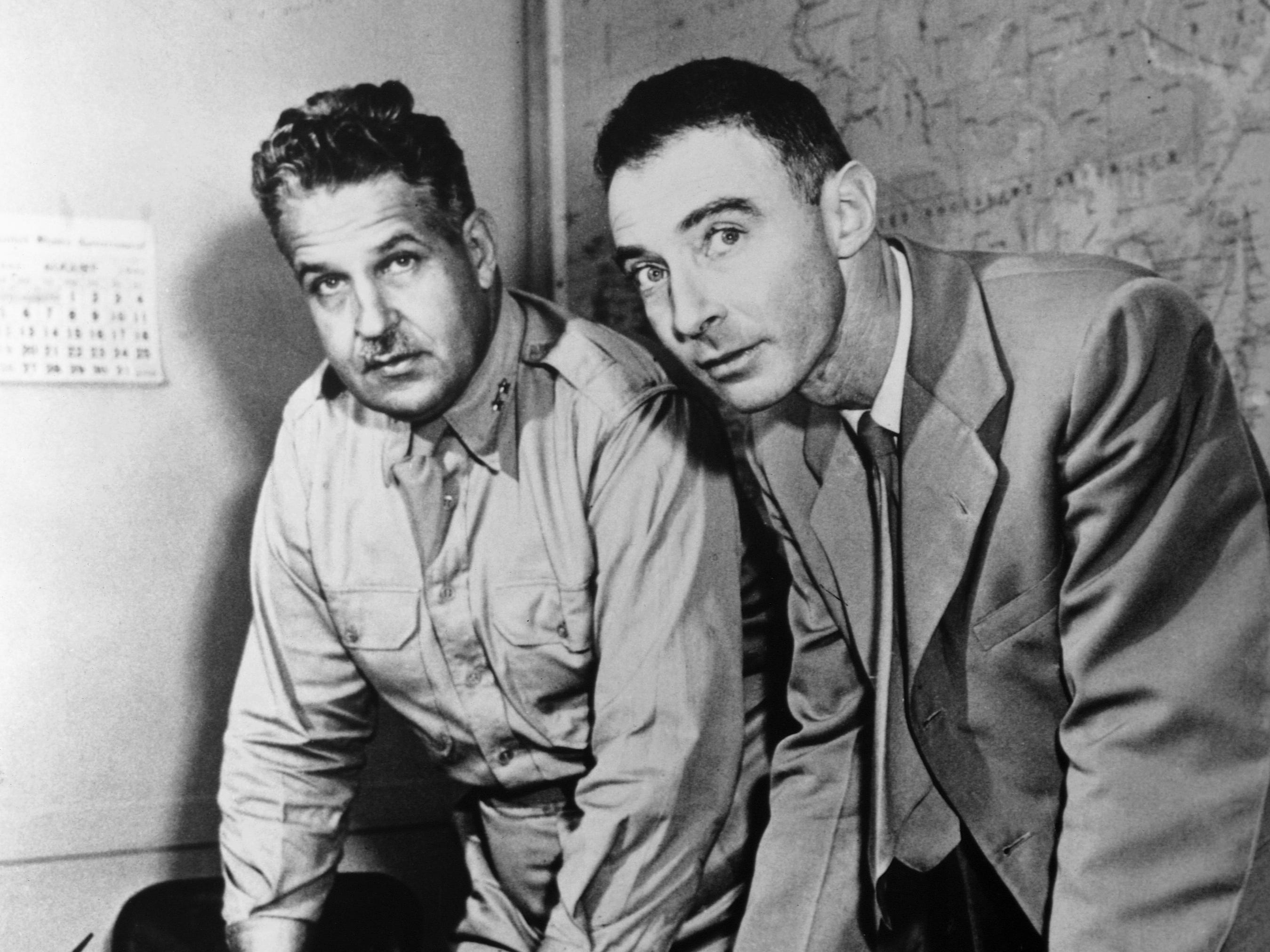 Schwarzweißfoto Leslie Groves in Uniform und Robert Oppenheimer im Anzug lehnen sich an einen Tisch vor einer Karte