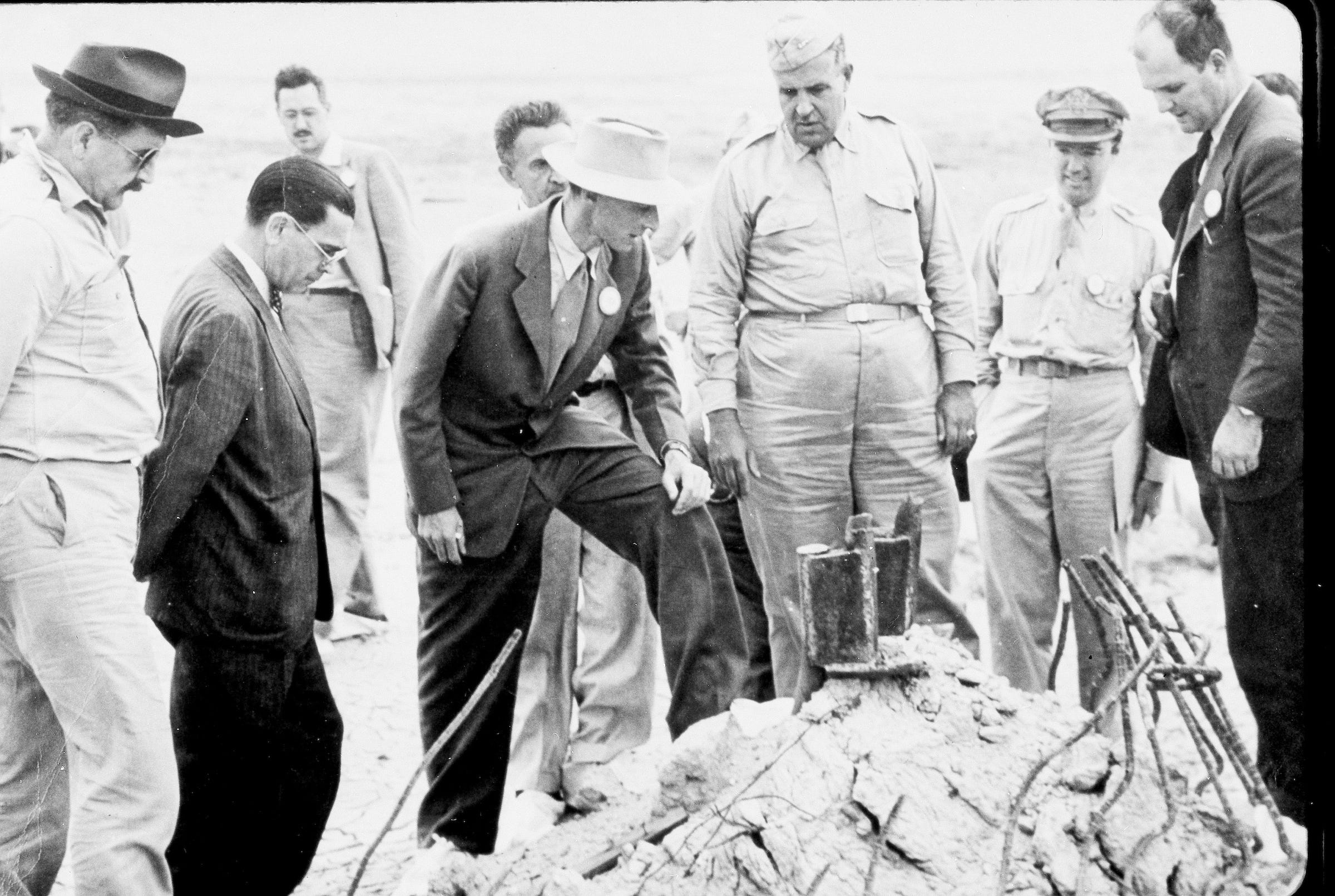 Neun Männer in Anzügen oder beigen Armeeuniformen stehen um einen Trümmerhaufen herum, Schwarz-Weiß-Foto