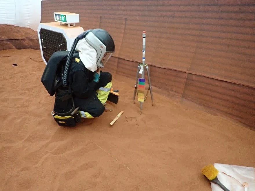 Eine Person kniet in einem schwarzen Raumanzug mit Helm und Sauerstoffrucksack nieder und blickt auf ein Stativ in einem mit rotem Sand gefüllten Raum