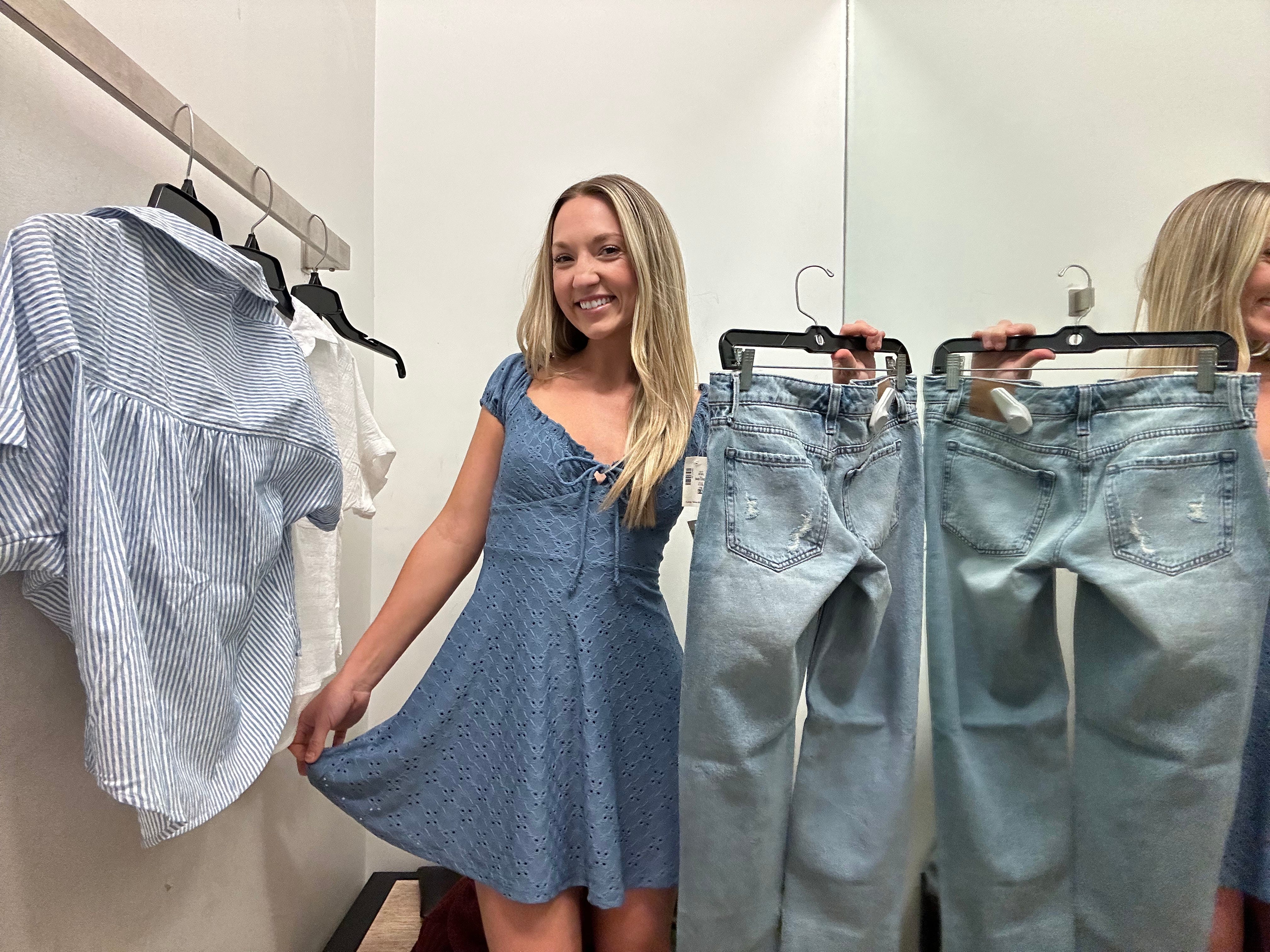 Danielle lächelt, während sie ein blaues Aéropostale-Kleid und eine Jeans in der Umkleidekabine trägt.  Links von ihr hängen ein paar Hemden auf Kleiderbügeln.