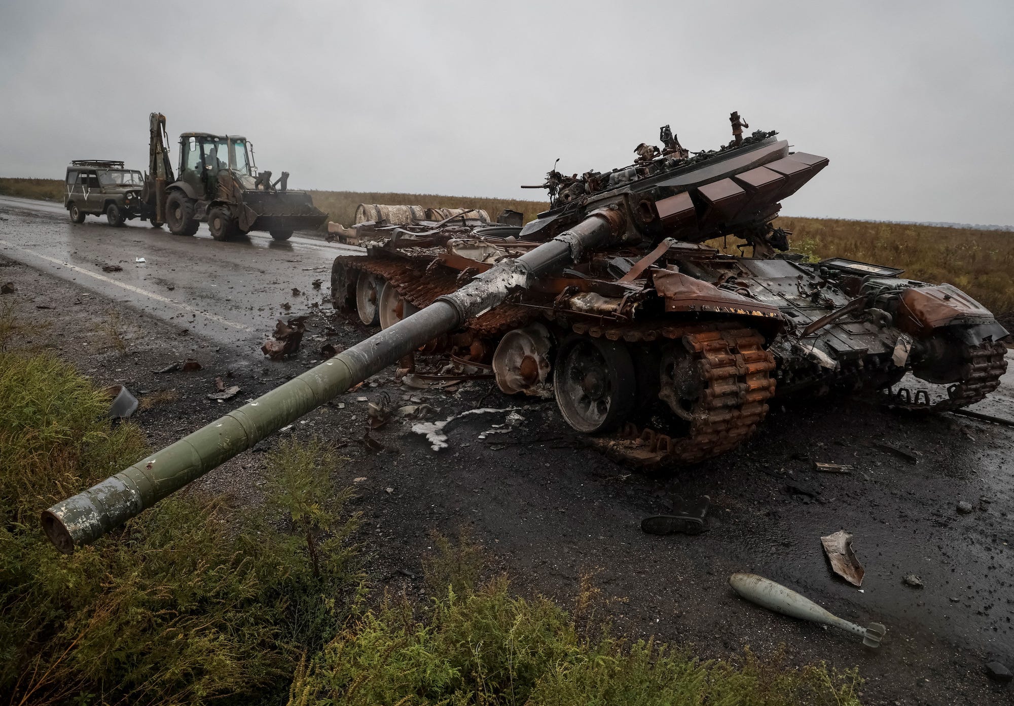Eine zerstörte russische Bräune auf einer Straße im Vordergrund, mit einem ukrainischen Soldaten, der auf einem Traktor fährt und ein Militärfahrzeug hinter sich herzieht, alles unter einem grauen Himmel