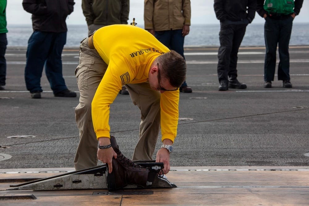 Lt.Cmdr.  Alex Cleaveland befestigt ein Paar Stiefel an einem Katapult, das vom Flugdeck eines Flugzeugträgers abgeschossen werden soll