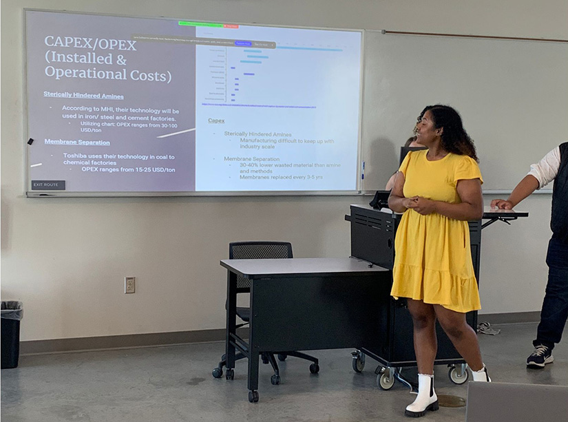Eine Schülerin in einem gelben Kleid steht vor einem Klassenzimmer und präsentiert eine Präsentation, während hinter ihr eine Folie auf einem Bildschirm läuft.