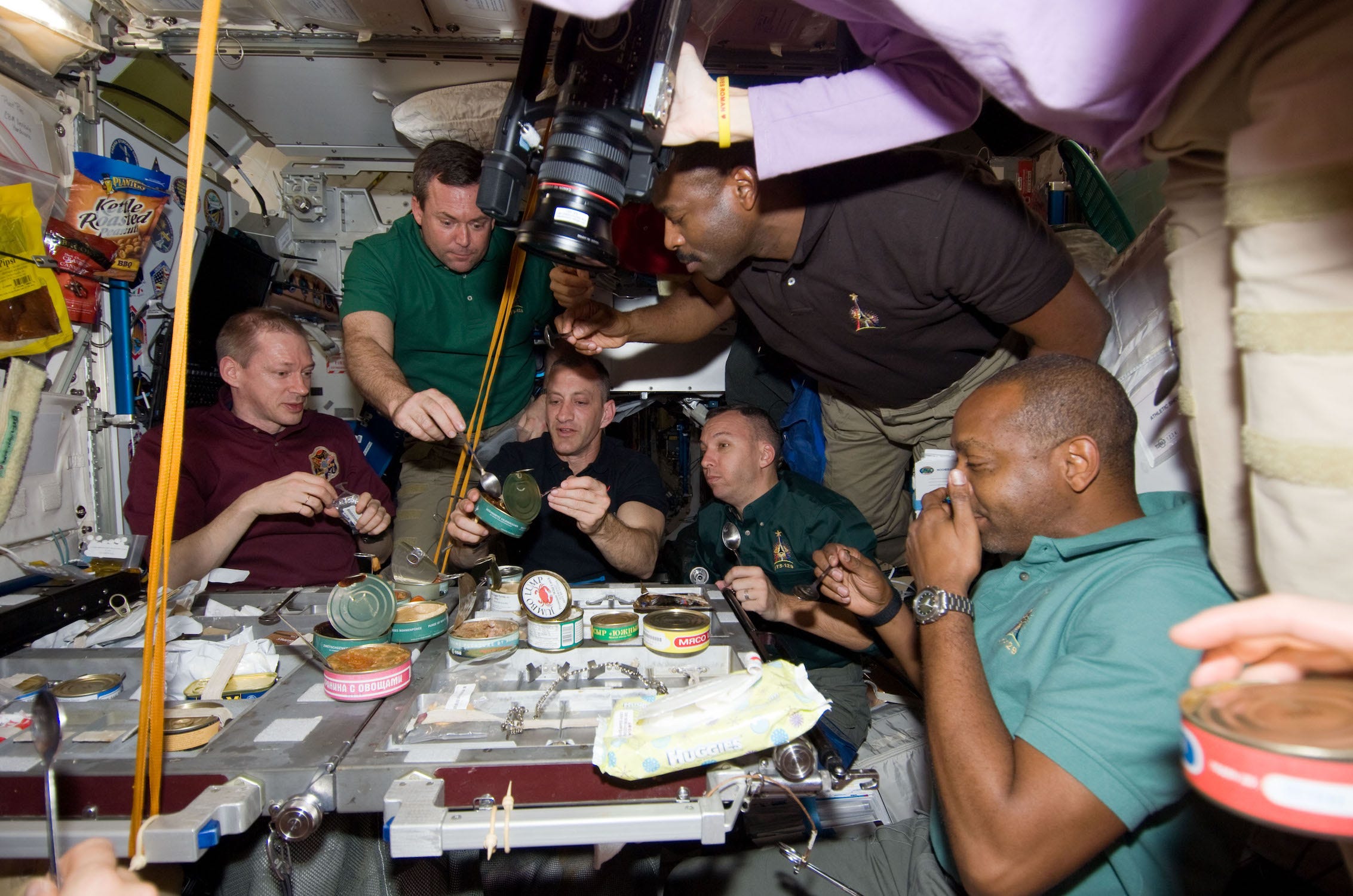 In der Internationalen Raumstation versammeln sich mehrere Menschen um einen Tisch, auf dem Dosen und andere Lebensmittel stehen