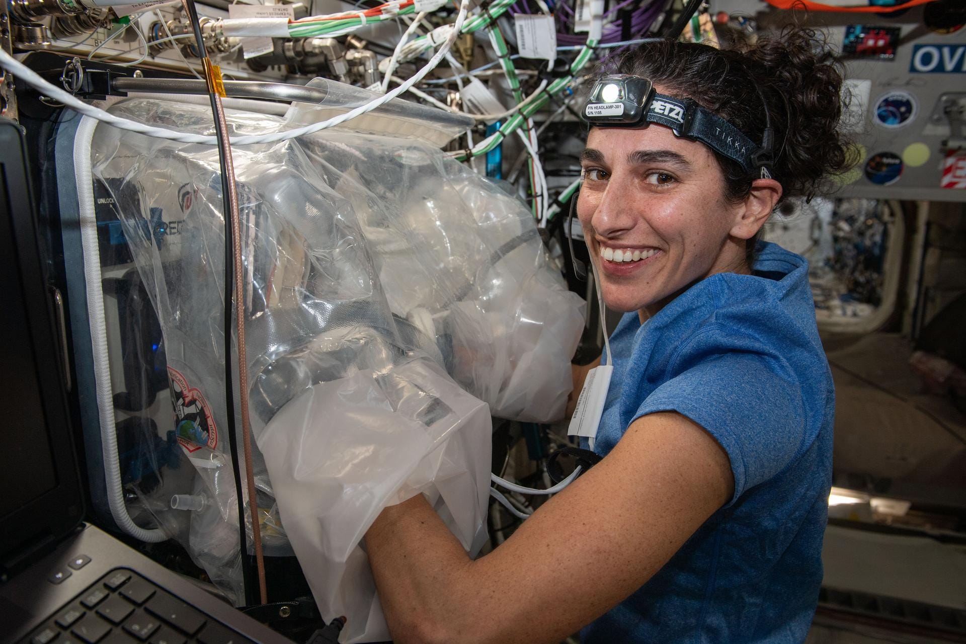Eine Astronautin mit eingeschaltetem Stirnband hat ihre Arme in einem großen Gerät mit Plastik darauf auf der Internationalen Raumstation