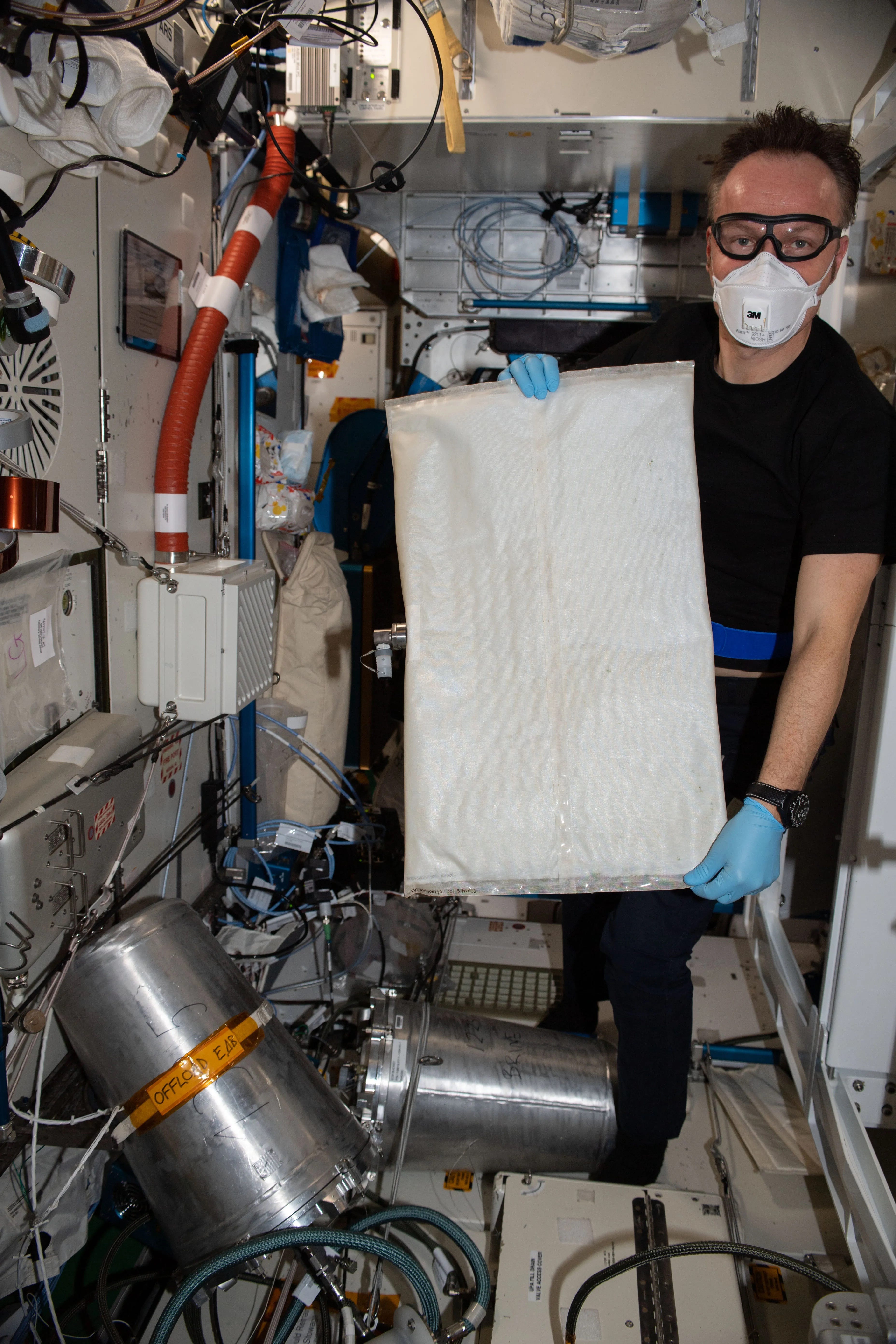 Astronaut in n95 und Schutzbrille und medizinischen Handschuhen hält ein großes, baumwollähnliches Rechteck in einem kleinen Raum innerhalb der Raumstation hoch