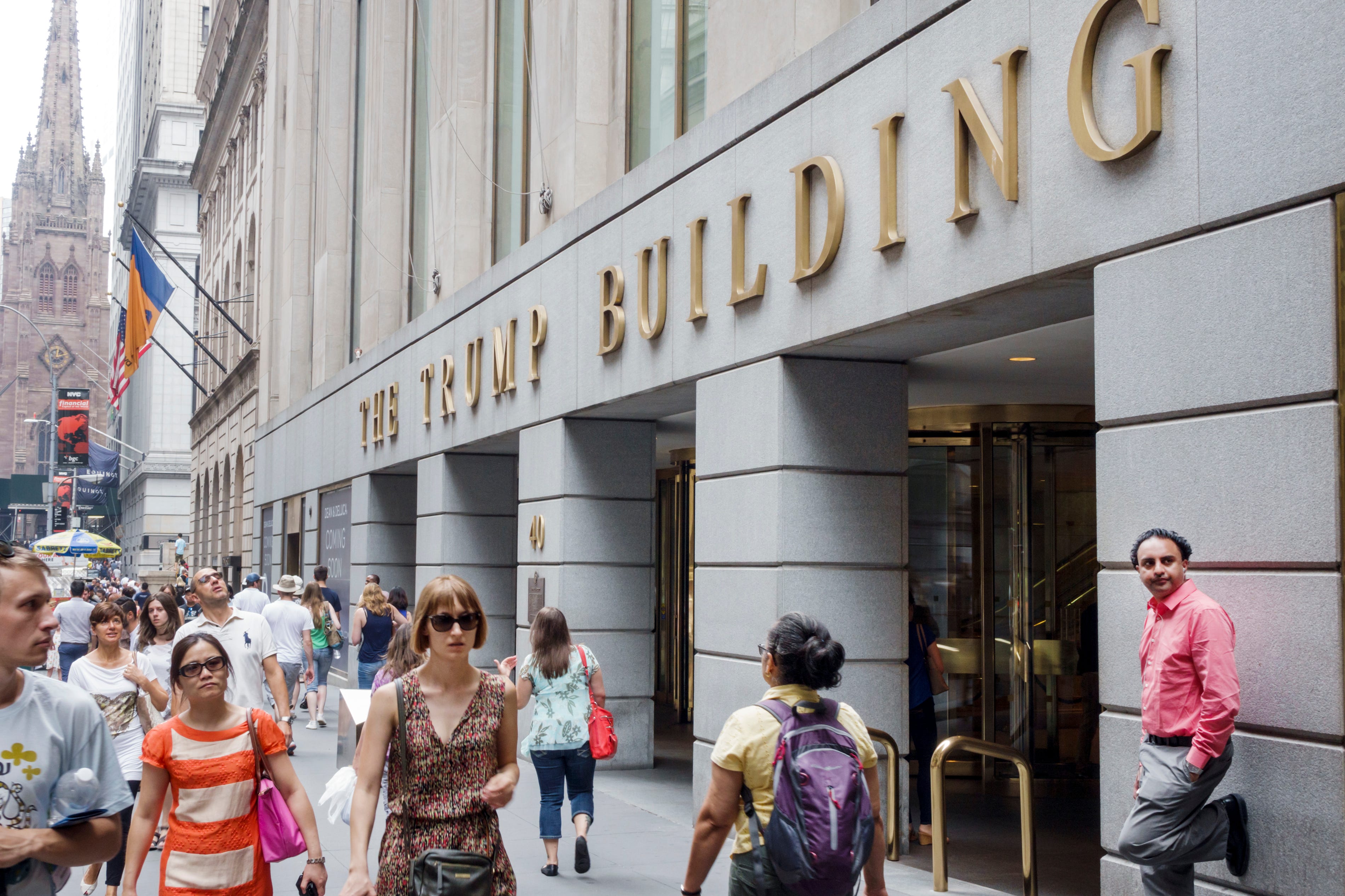 Eine Menge New Yorker geht an der grauen Fassade von 40 Wall Street vorbei, wo in goldenen Buchstaben „The Trump Building“ steht