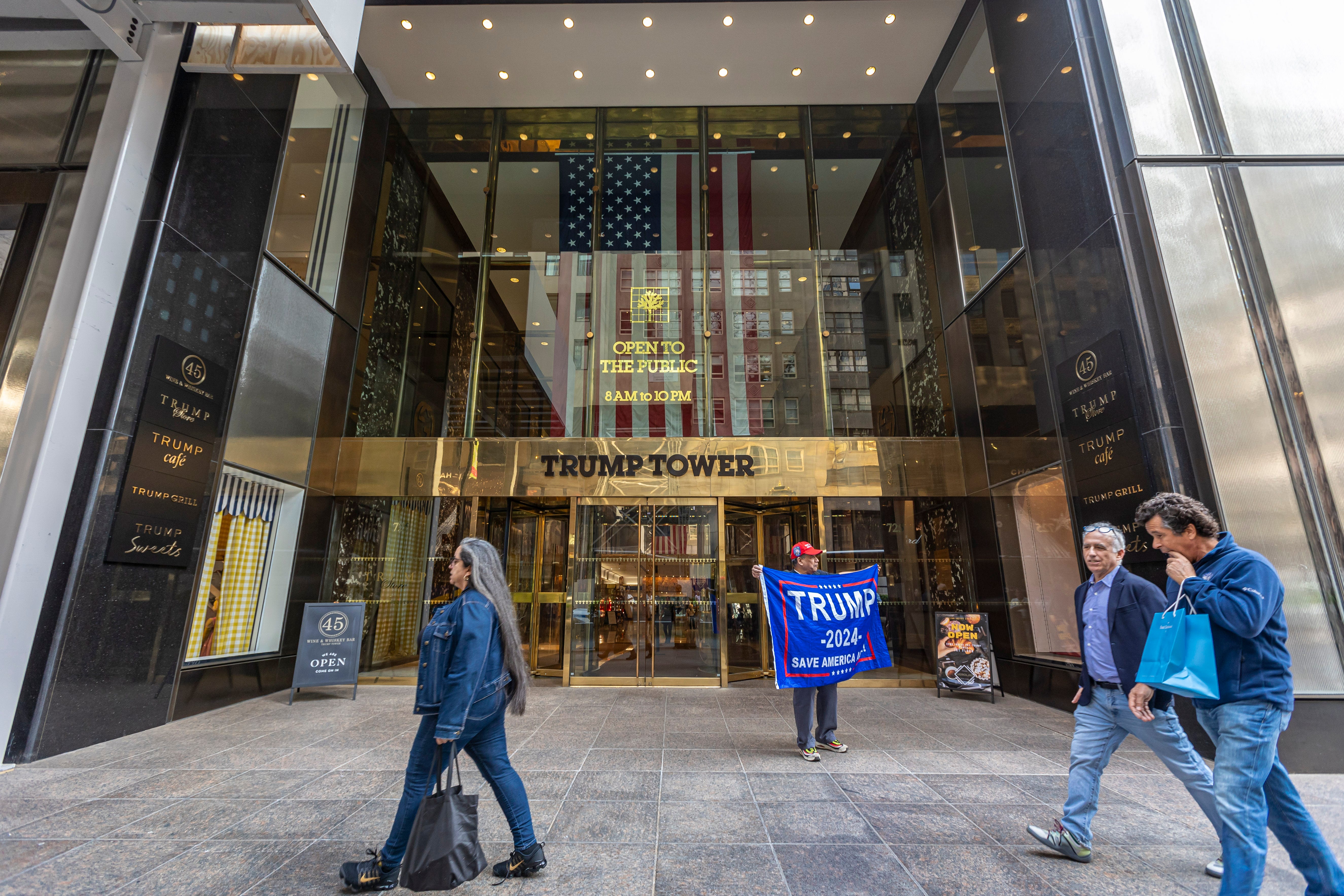 Ein Mann hält eine „Trump 2024“-Flagge vor die vergoldete Glasfassade der Lobby des Trump Tower
