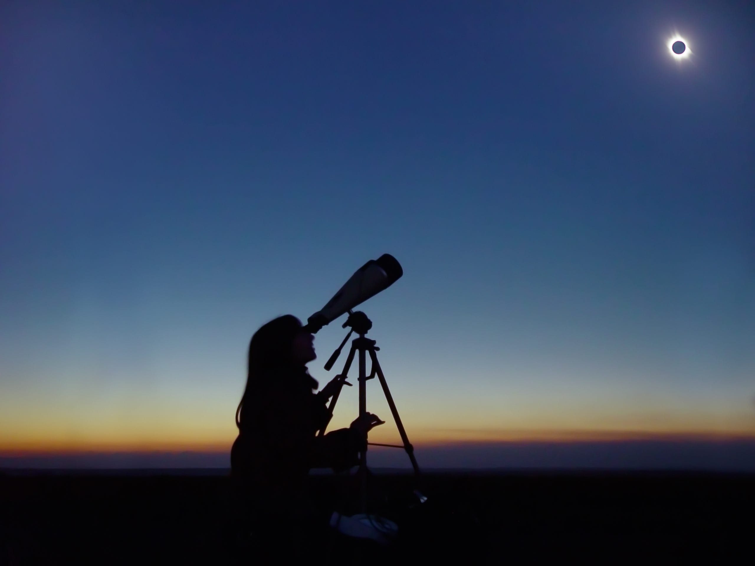 Eine Person betrachtet eine totale Sonnenfinsternis durch ein Teleskop mit Sonnenuntergangsfarben am Horizont hinter ihr.