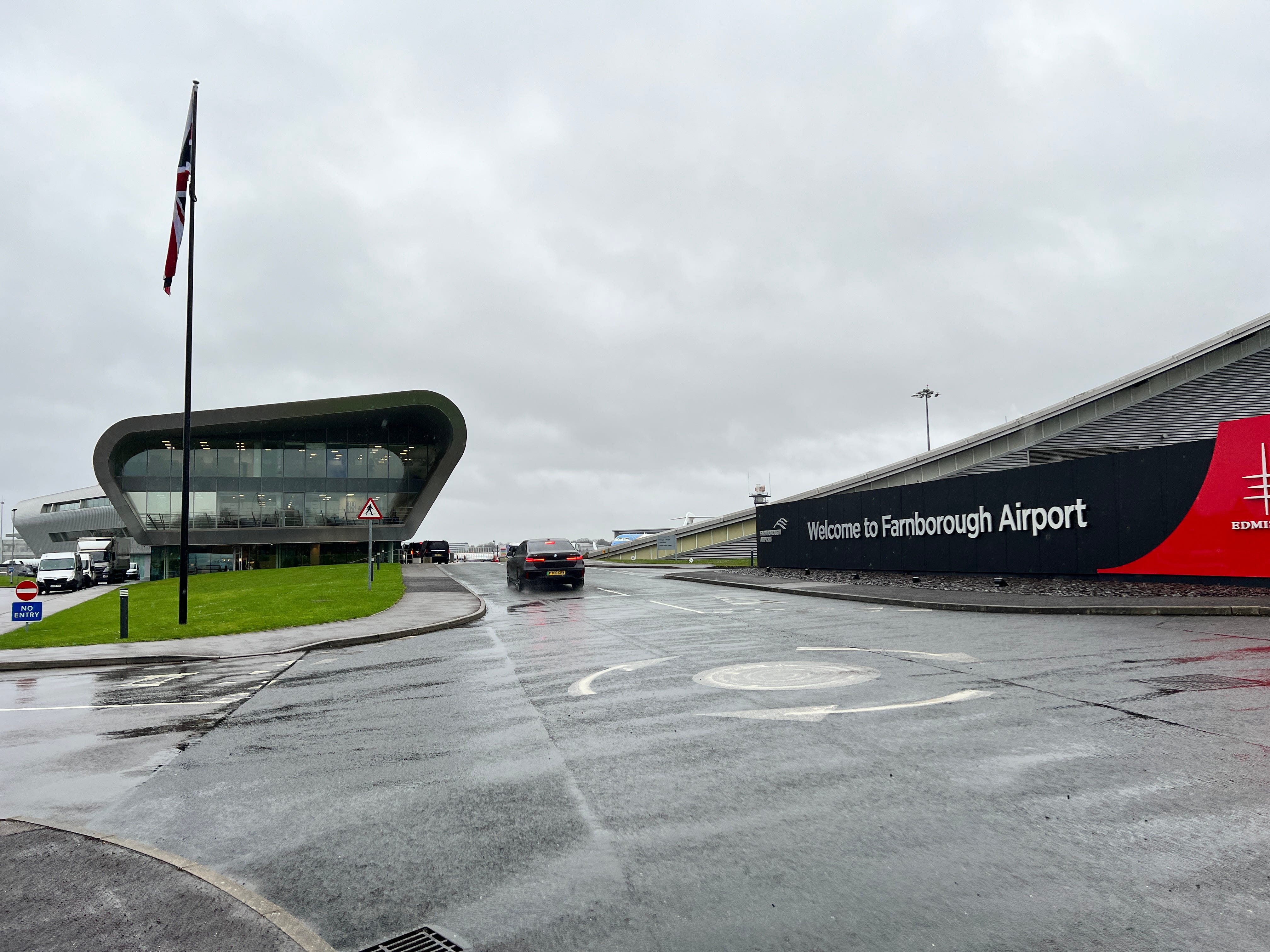 Der Eingang zum Flughafen Farnborough zeigt ein Schild und ein modernes Terminalgebäude, während ein Auto vorbeifährt.