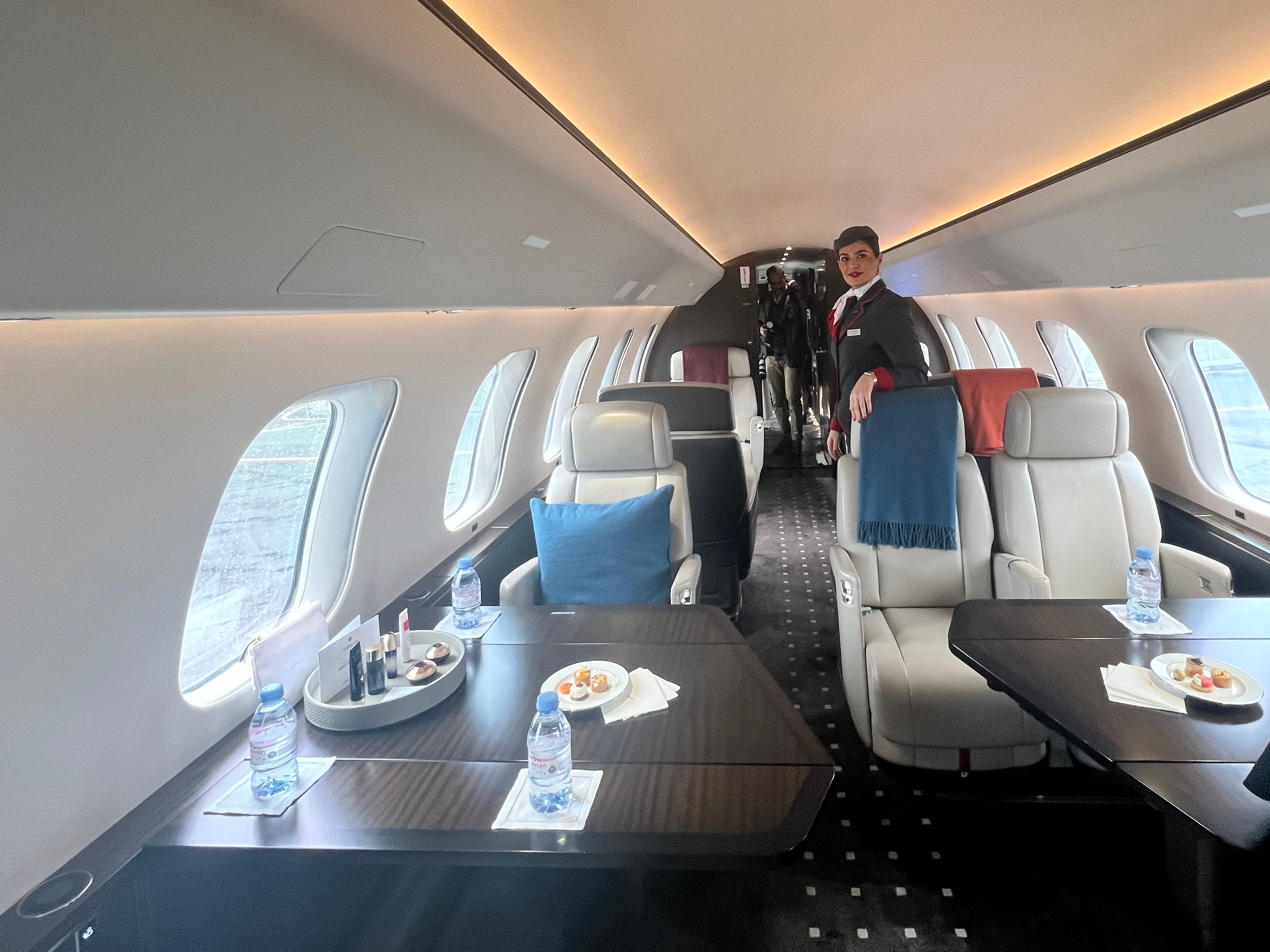 Eine Flugbegleiterin steht im Hauptsitz-/Essbereich an Bord einer VistaJet Bombardier Global 7500 am Flughafen Farnborough, wo neun cremefarbene Ledersitze zu sehen sind.