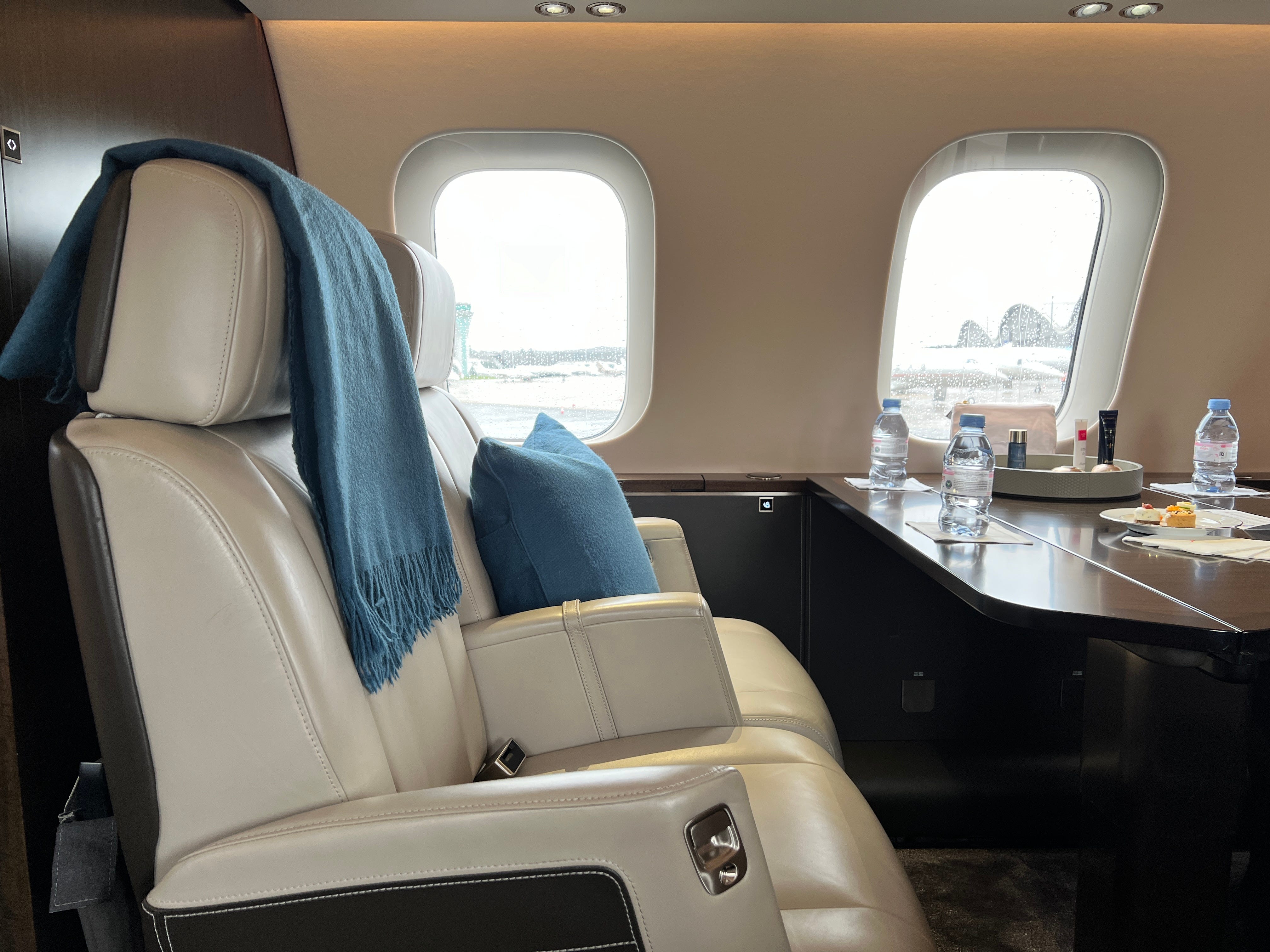 Zwei große cremefarbene Ledersitze mit blauer Decke und Kissen neben einem Tisch mit Wasserflaschen an Bord eines VistaJet Bombardier Global 7500