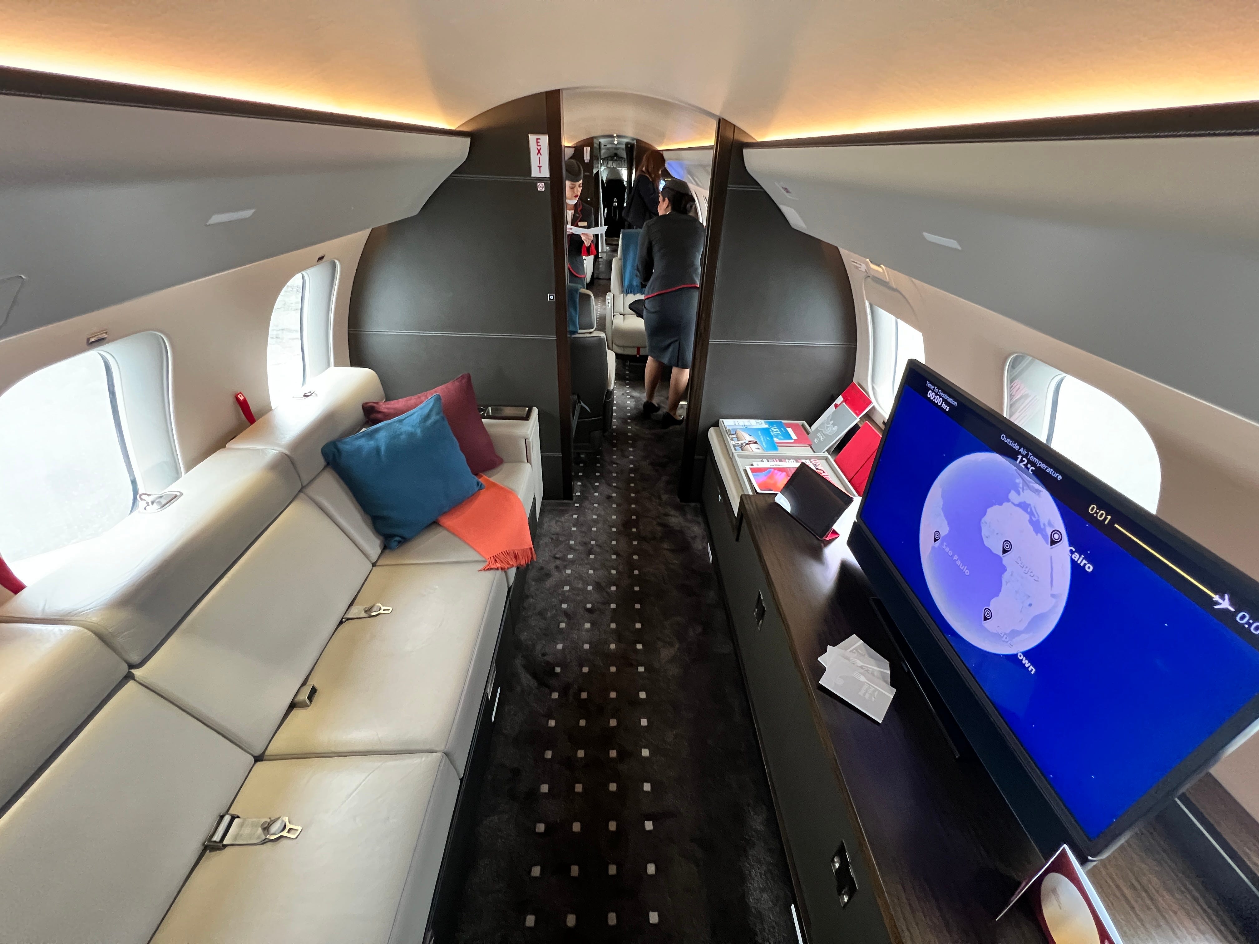 Der Fernseh-/Wohnbereich eines VistaJet Bombardier Global 7500 mit einem langen Sofa sowie Kissen und Literatur