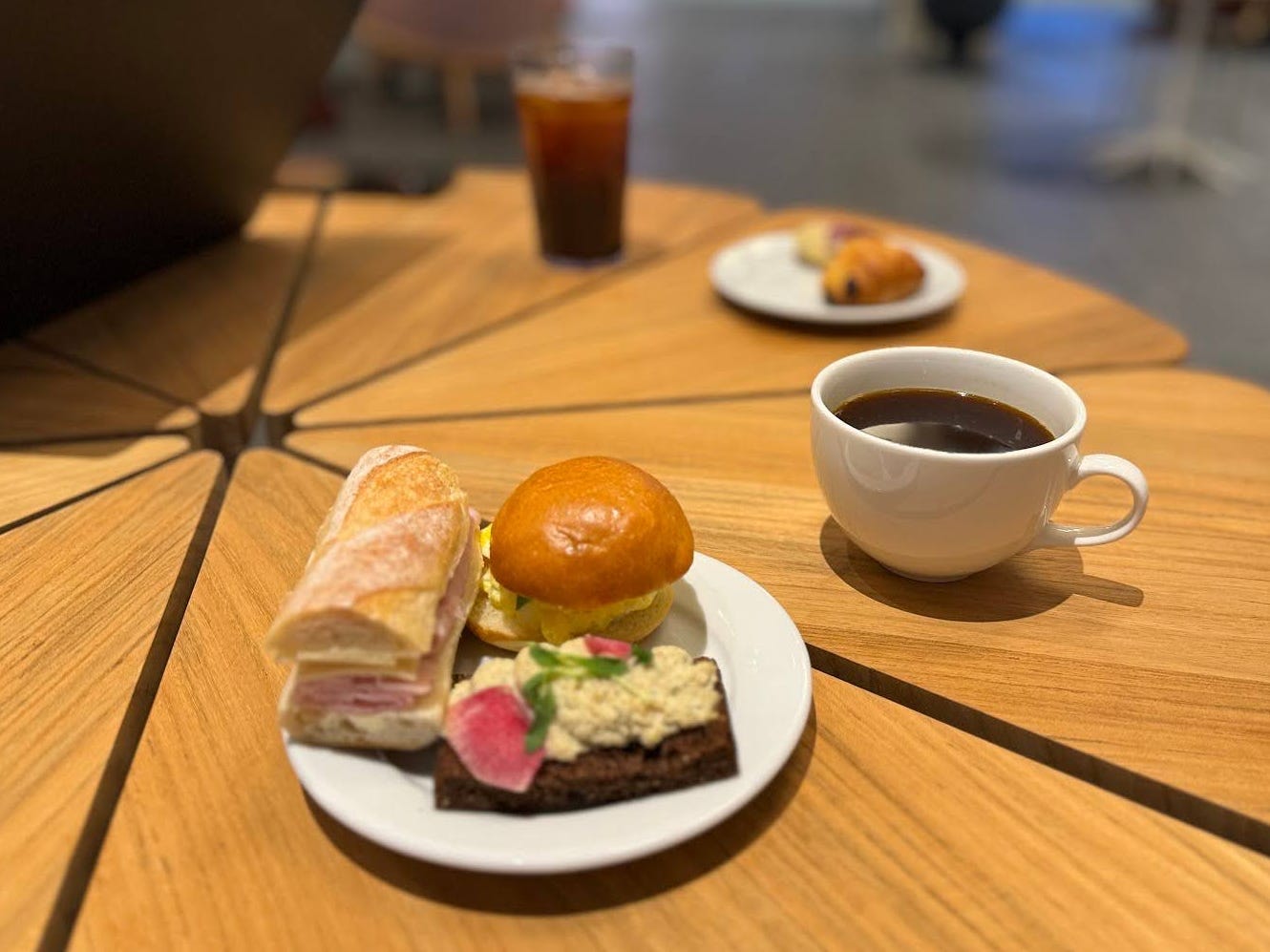 Ein Americano in einer Tasse und ein Mini-Eiersalat-Sandwich, ein Mini-Schinken-Schweizer-Sandwich und eine geräucherte Seeforellen-Tartine auf einem Teller.
