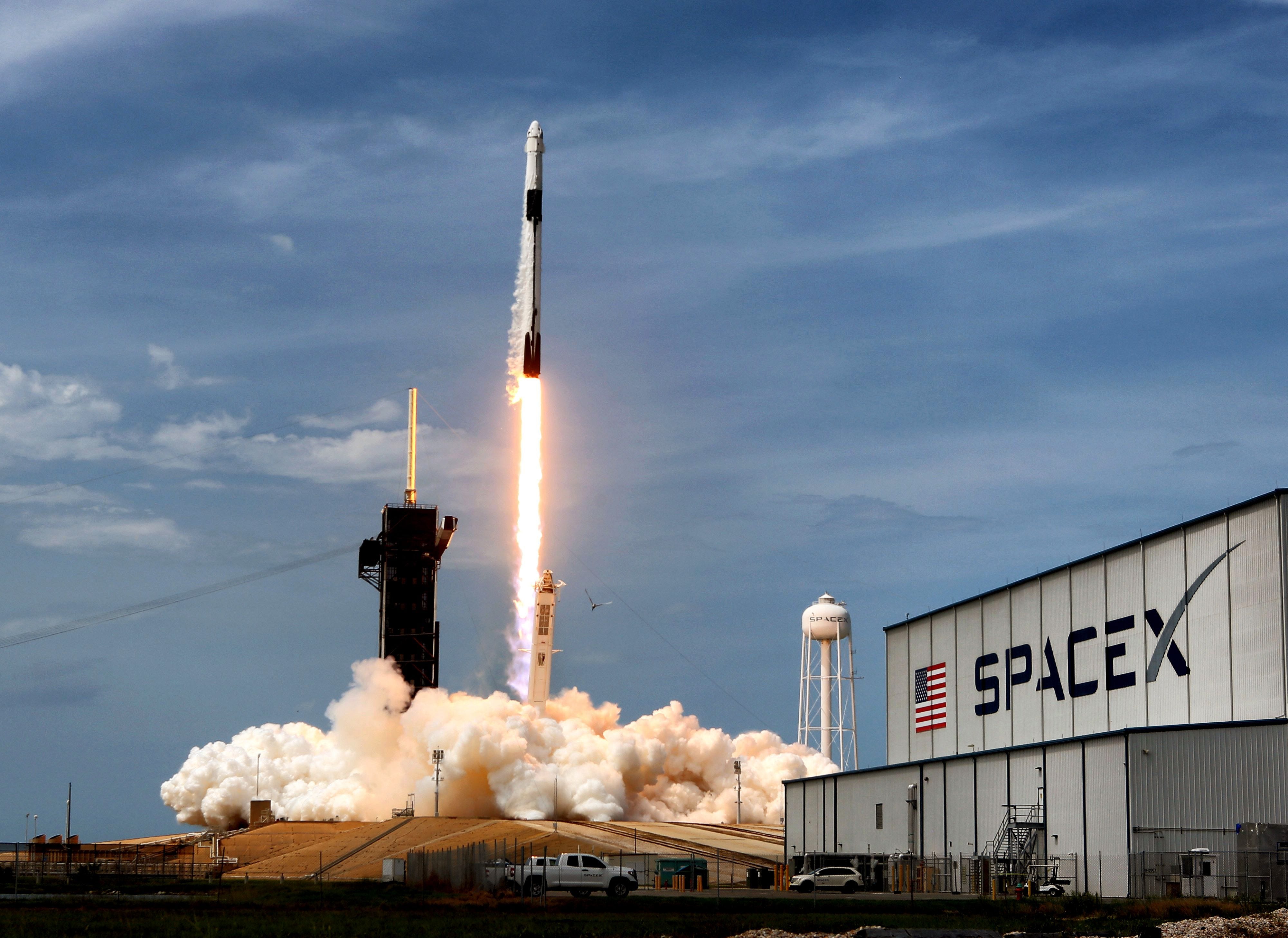 Eine SpaceX Falcon 9-Rakete startet vor einem Abendhimmel, im Vordergrund ist eine Fabrik mit SpaceX-Logo zu sehen.