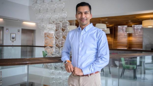 Aron D'Souza lächelt, während er neben einer Glasskulptur in einem Großraumbüro posiert