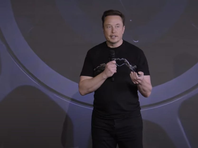 Elon Musk spricht in ein Mikrofon, während er eine Brille in der Hand hält.