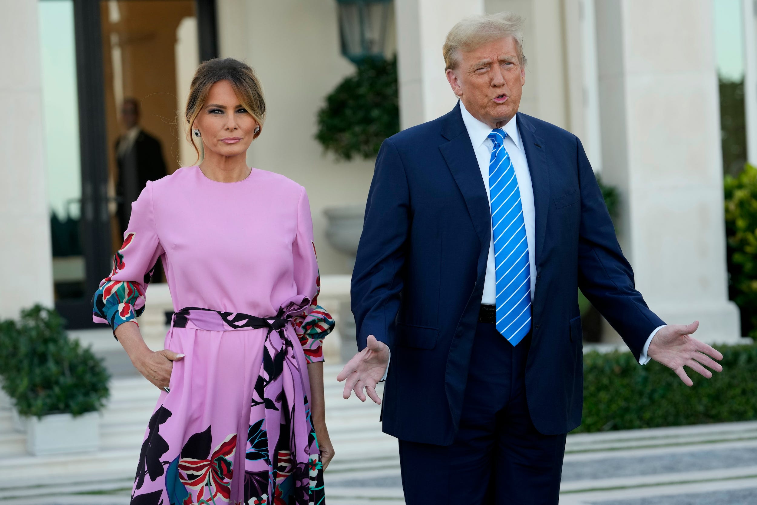 Melania Trump in einem rosa Kleid mit langen Ärmeln und Donald Trump in einem blauen Anzug und einer gestreiften Krawatte sprechen mit Reportern vor einer weißen Villa