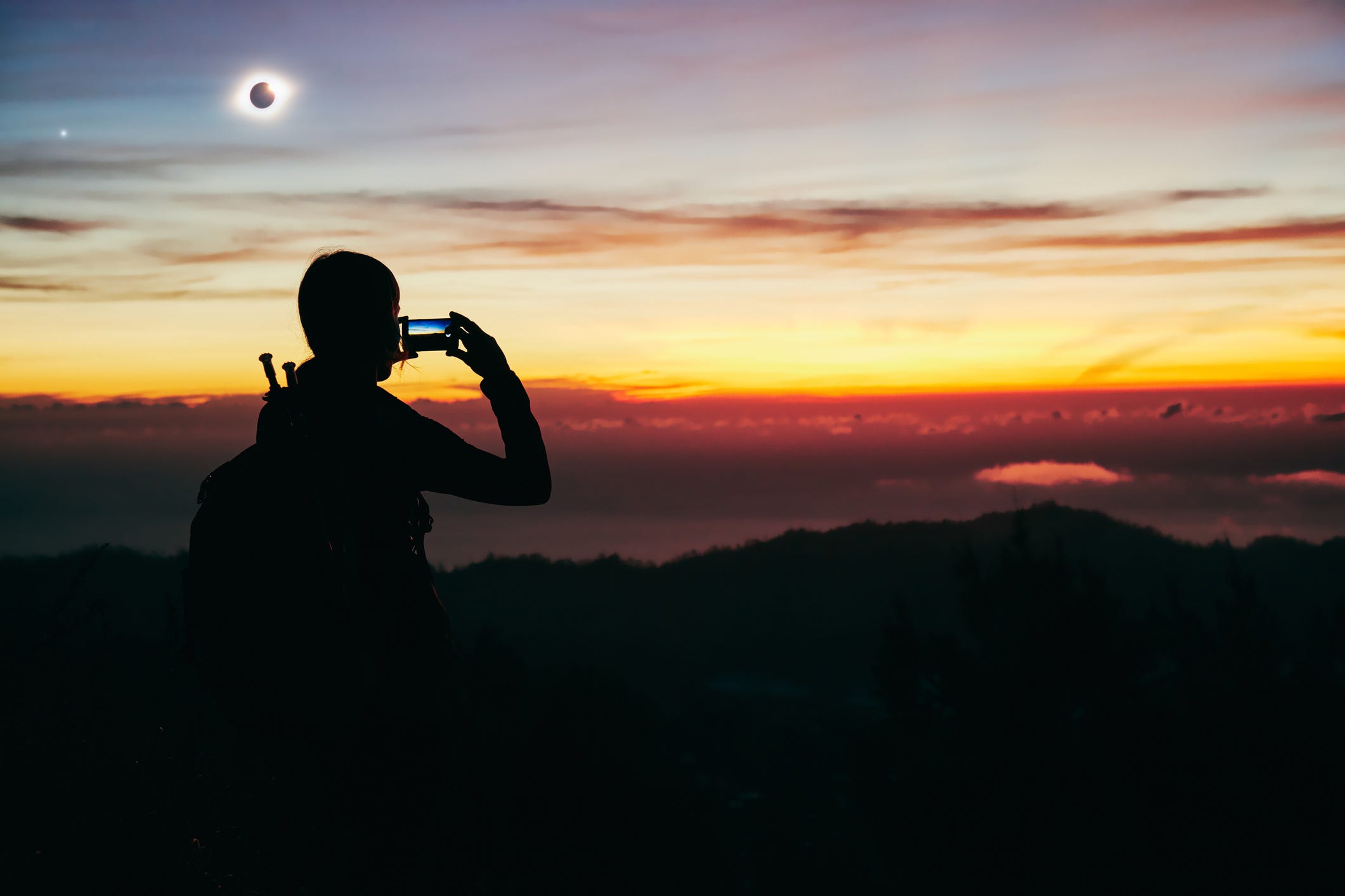 Die Silhouette einer Person, die mit ihrem iPhone ein Foto des sonnenuntergangsähnlichen Horizonts macht, mit einer totalen Sonnenfinsternis am Himmel darüber