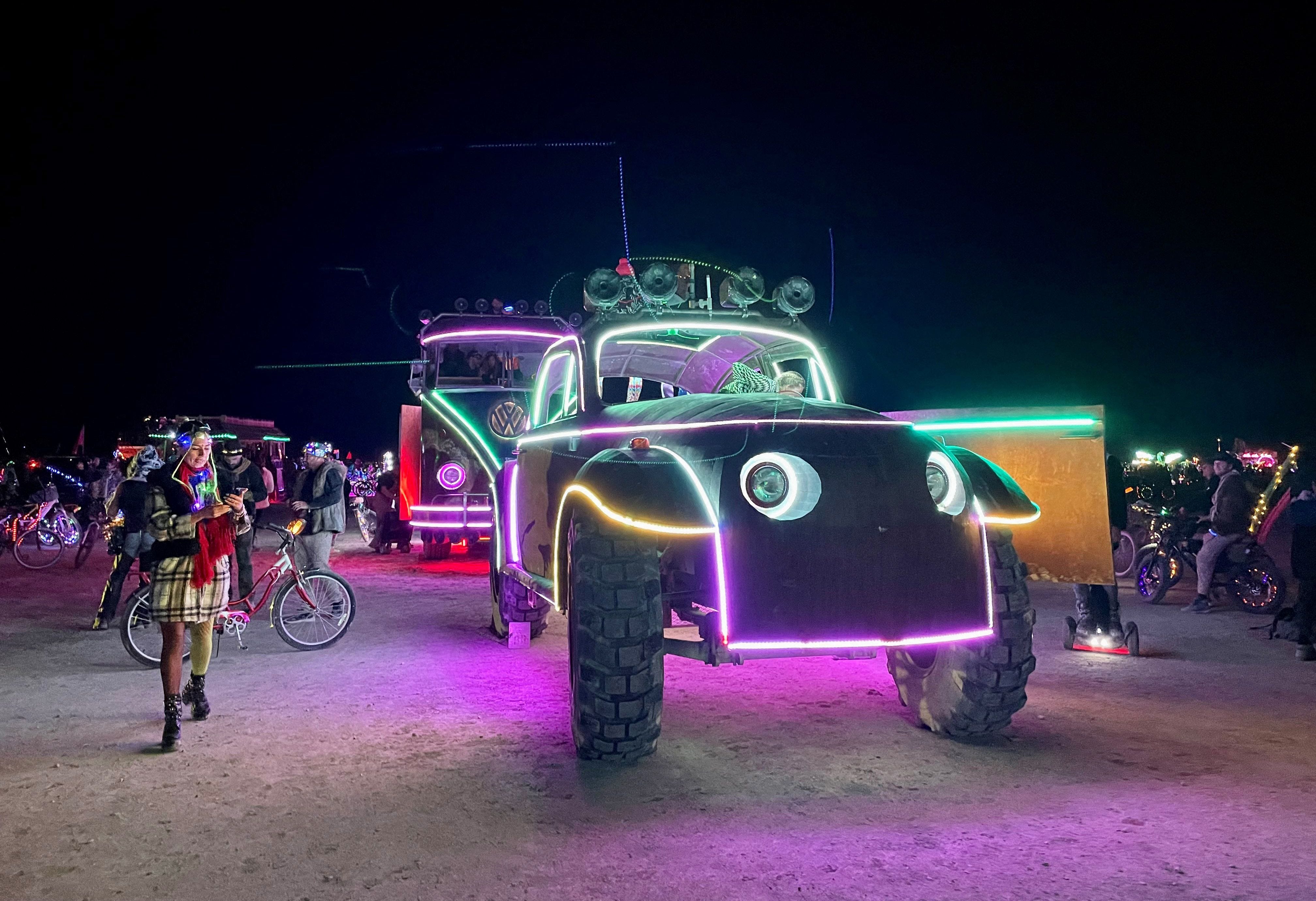 Eine Reihe von mit Neonlichtern geschmückten Fahrzeugen fahren durch die Playa des Burning Man, umgeben von Teilnehmern.