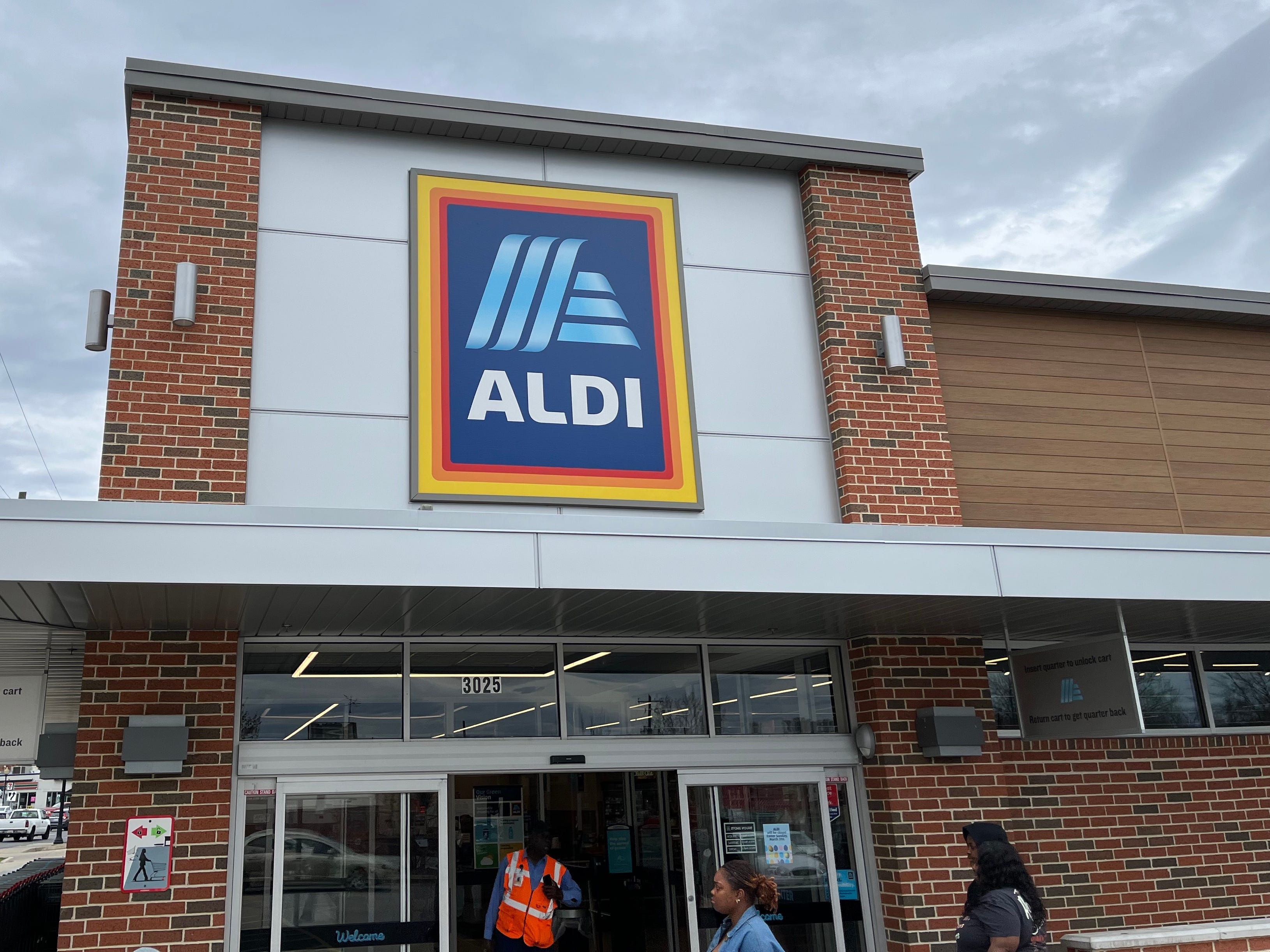 An einem bewölkten Tag in Hyattsville, Maryland, gehen Menschen in ein Aldi-Lebensmittelgeschäft ein und aus.  Über dem Eingang hängt das blau-weiße Aldi-Logo auf einem Schild, links vom Eingang steht eine Reihe Einkaufswagen.