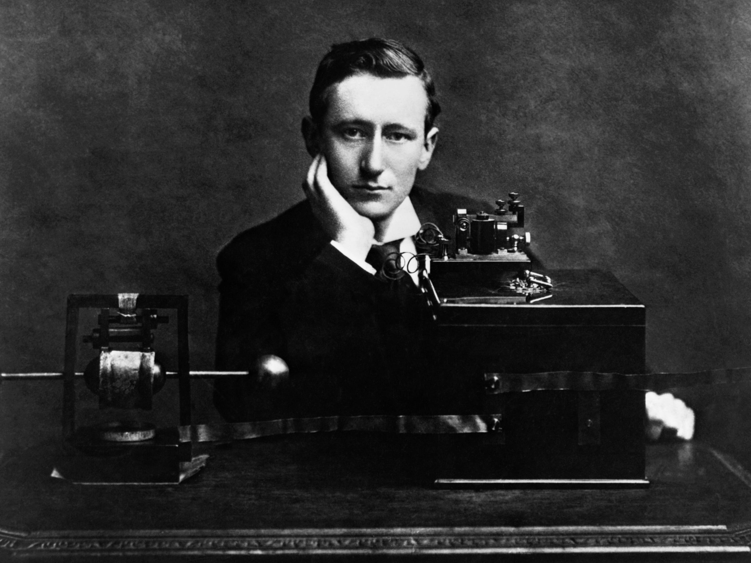 Gugliemo Marconi sitzt mit der Hand unter dem Kinn an einem Schreibtisch mit seinem elektrischen Funkgerät