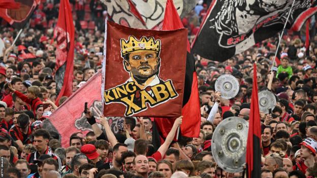 Leverkusen-Fans feiern auf dem Spielfeld mit einem Banner mit der Aufschrift „König Xabi“ als Hommage an Trainer Xabi Alonso