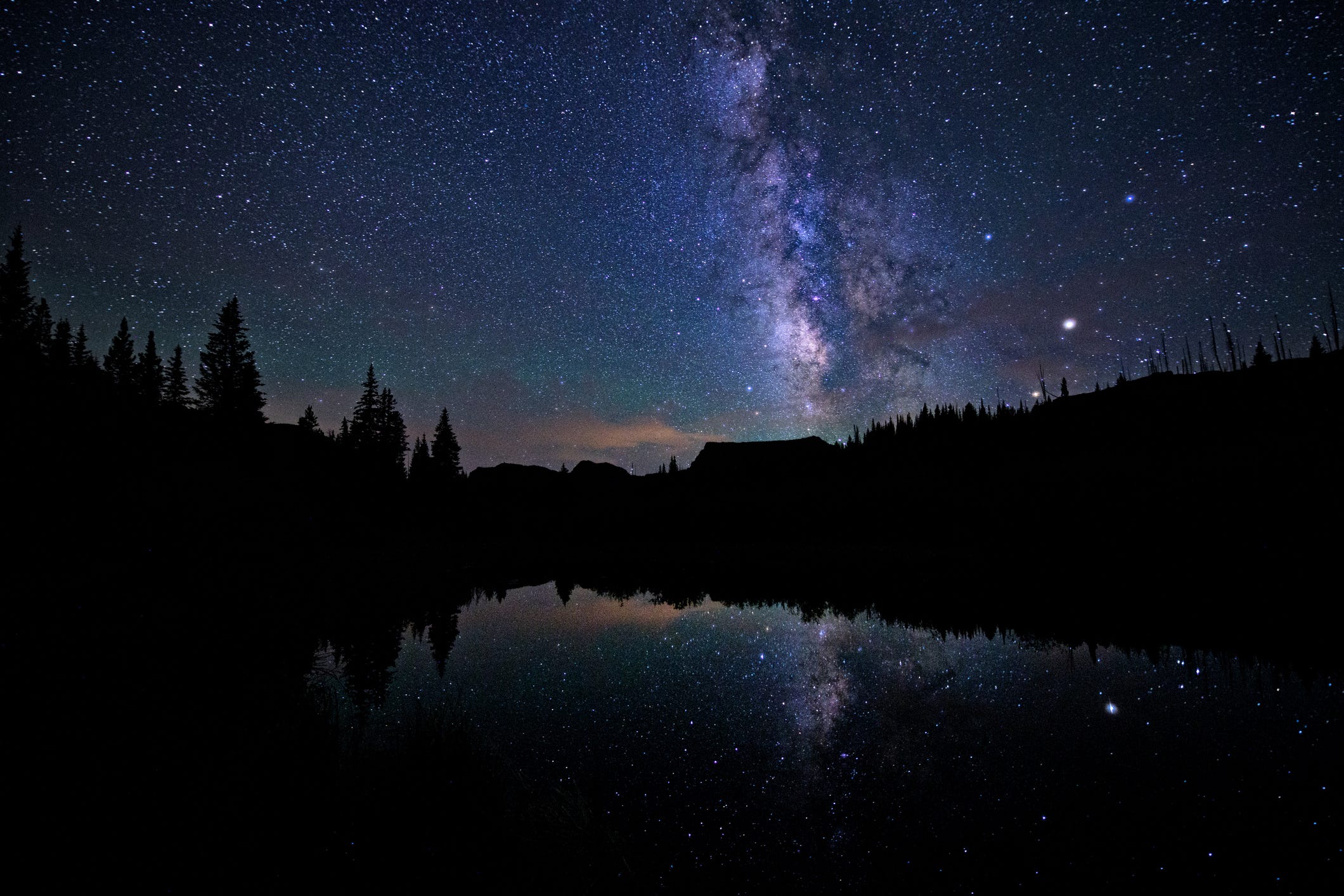 Ein sternenklarer Nachthimmel mit Milchstraße über einem von Wald umgebenen Teich.