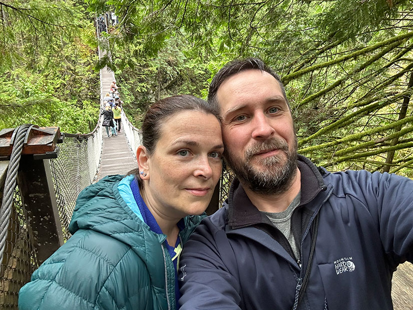 Ein Selfie von zwei Menschen auf einer Hängebrücke, hinter ihnen gehen Menschen auf der Brücke und Bäume umgeben sie.