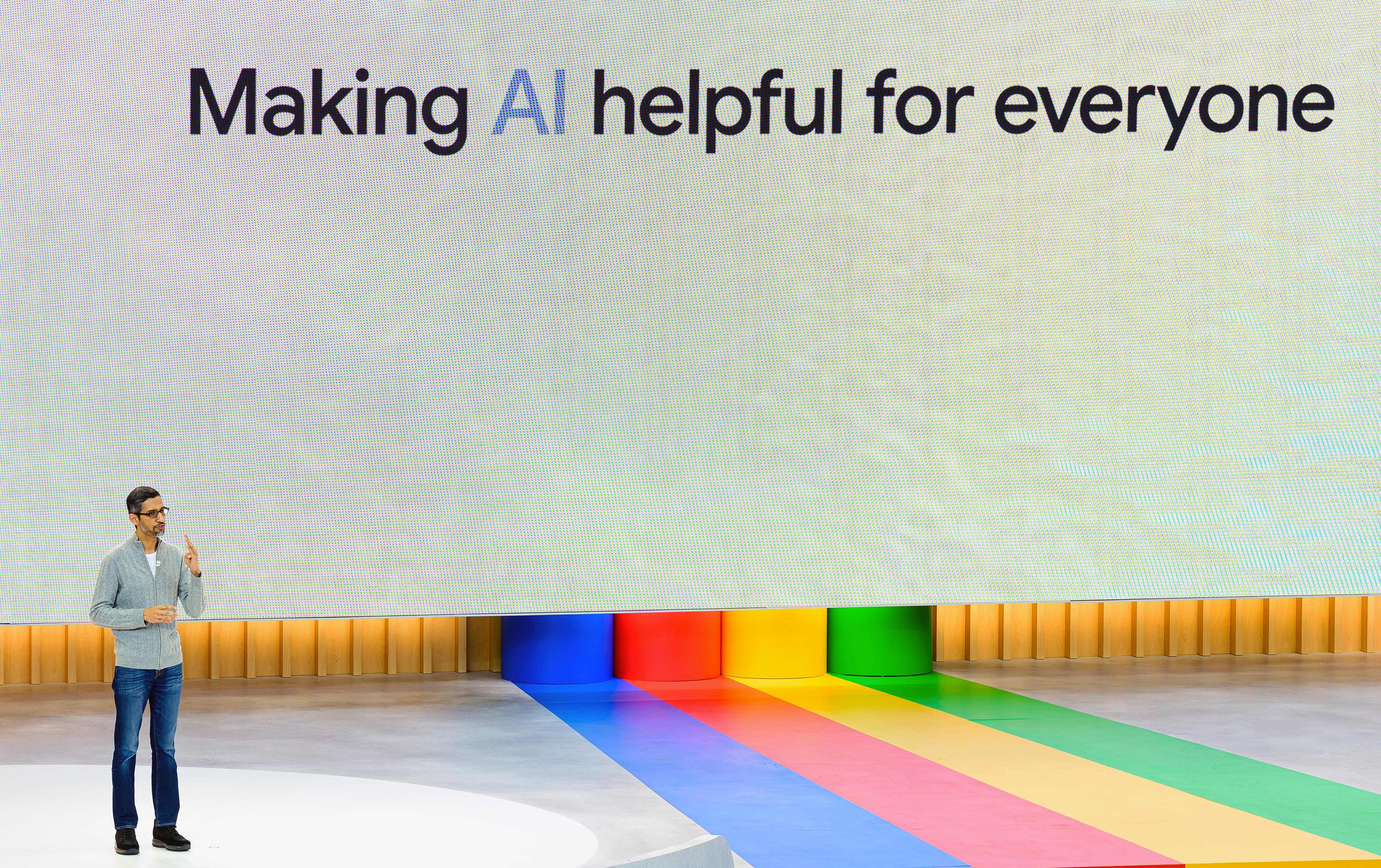 Google-Chef Sundar Pichai hält auf einer Bühne vor einem Bildschirm eine Rede mit der Aufschrift „KI für alle hilfreich machen“.