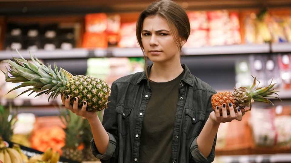 Young woman skeptically holds up a pineapple in the supermarket