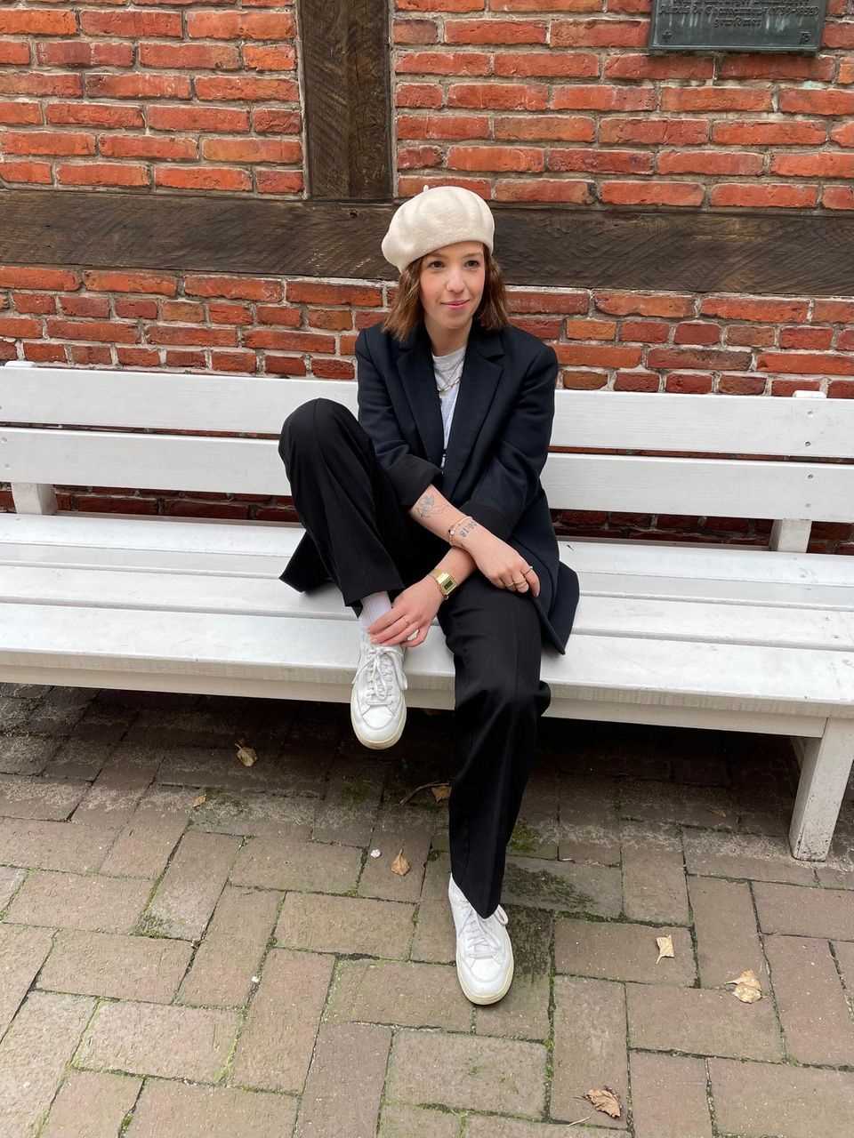 Brunette woman with black suit and white sneakers and beige cap on a white bench