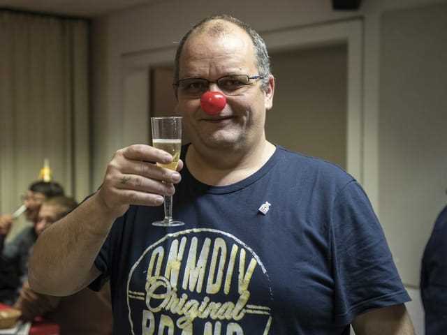 Ein Mann hält ein Glas Sekt in der Hand und trägt eine rote Aufsteck-Nase. 