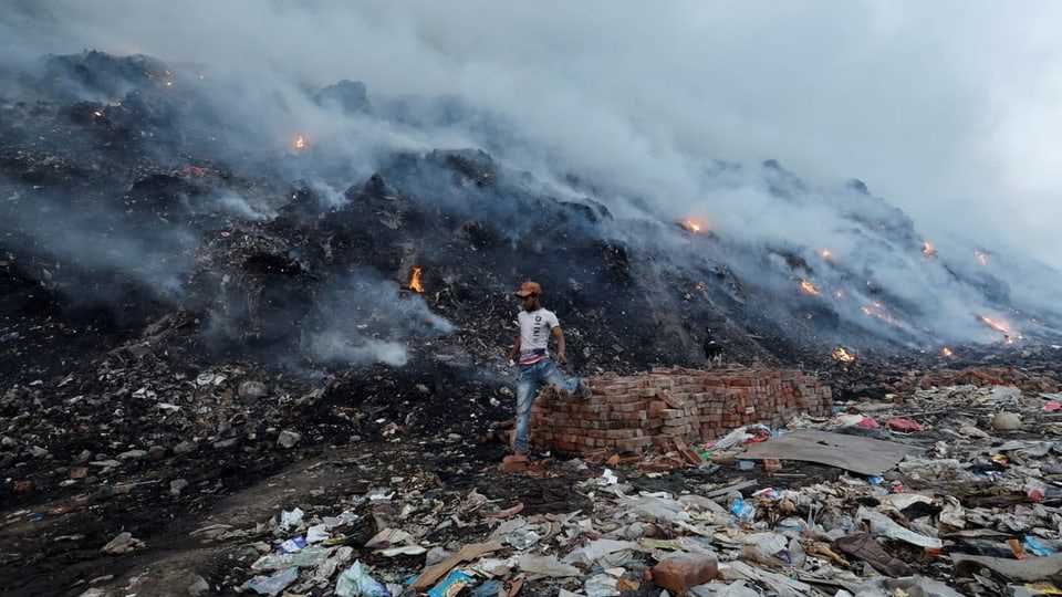 A man walks past a slightly burning garbage dump.