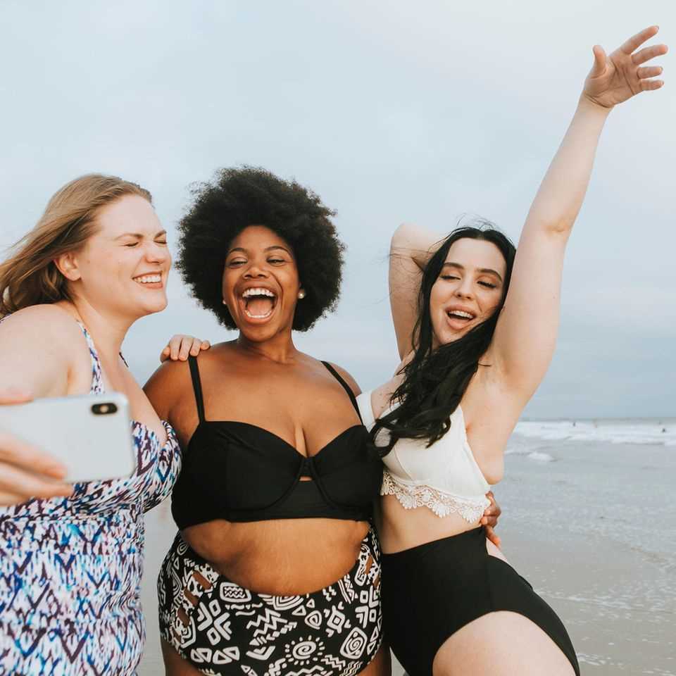 Shape Bikini, three laughing women in swimwear on the beach taking a selfie