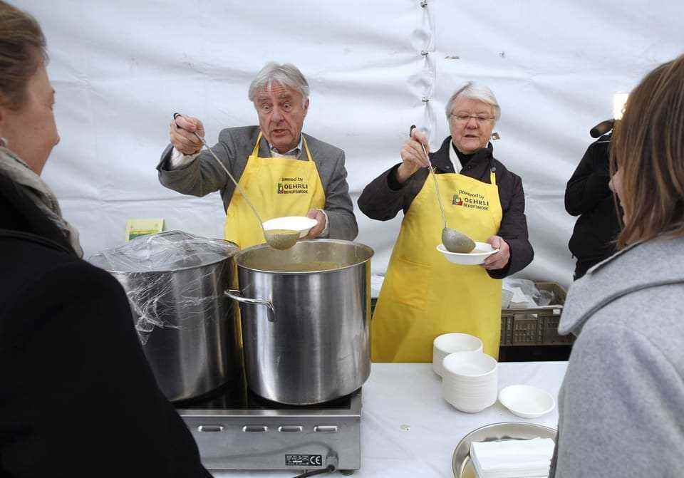 A man with white hair and a woman with white hair are scooping soup in a tent, wearing yellow aprons.