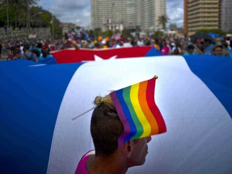 Man in front of a giant Cuban flag, has a rainbow flag in his hair.