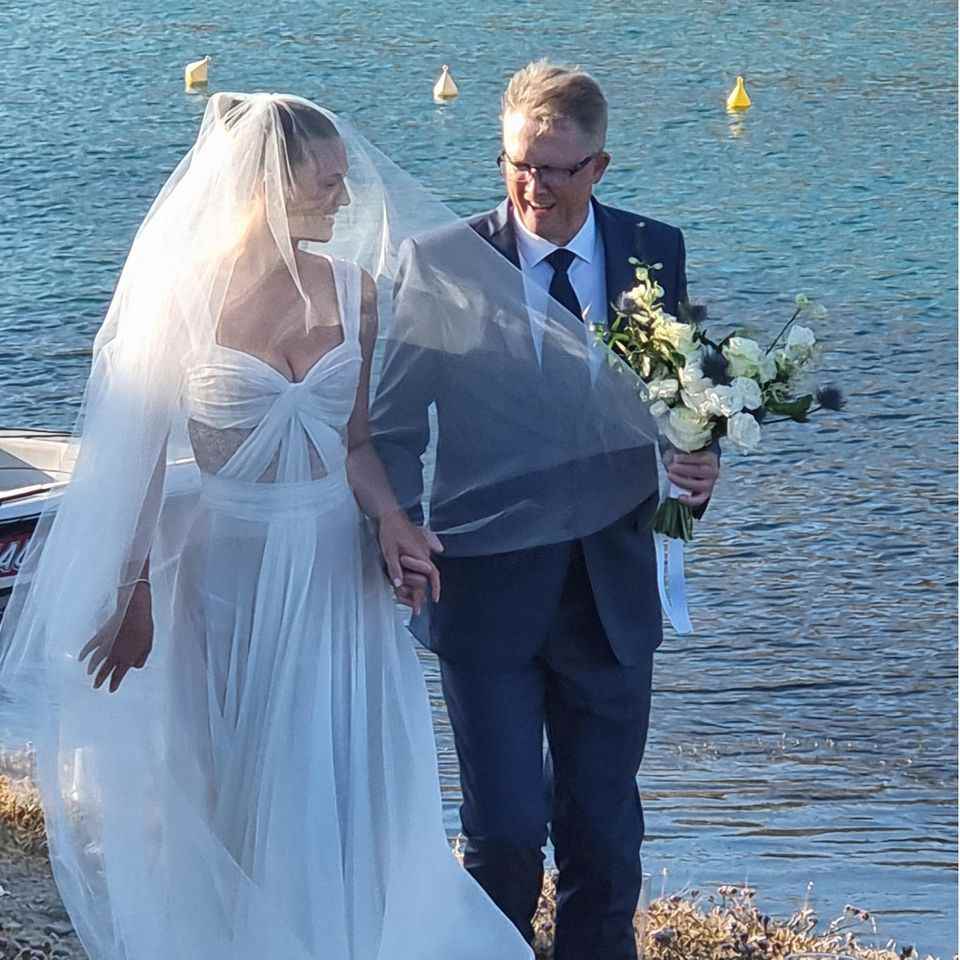 Her father leads Toni to the altar in front of the romantic dream scenery of Paros.  The dress, tailor-made for Toni, impresses with its flowing, slightly transparent tulle fabric and knotted look.  The bride wears natural make-up. 