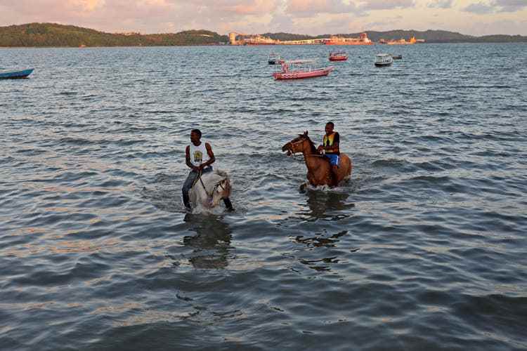 Two men ride their horses in the sea in front of the Quilombo Banairas on Ilha de Mare.
