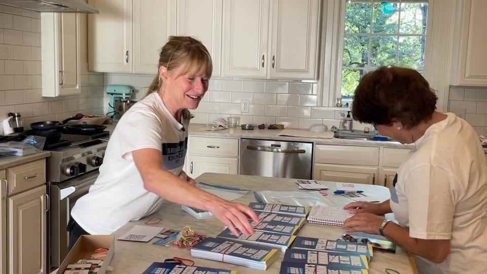 Two women bundle flyers in a kitchen.