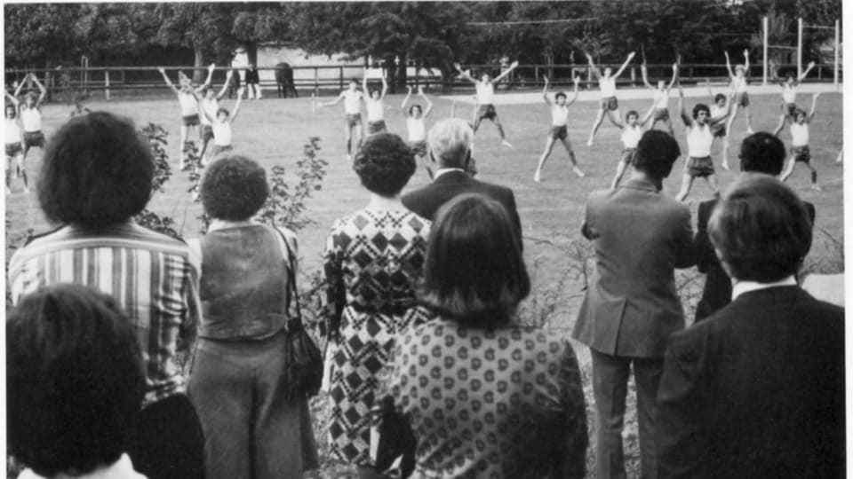 Children do gymnastics on a meadow and are watched by adults.