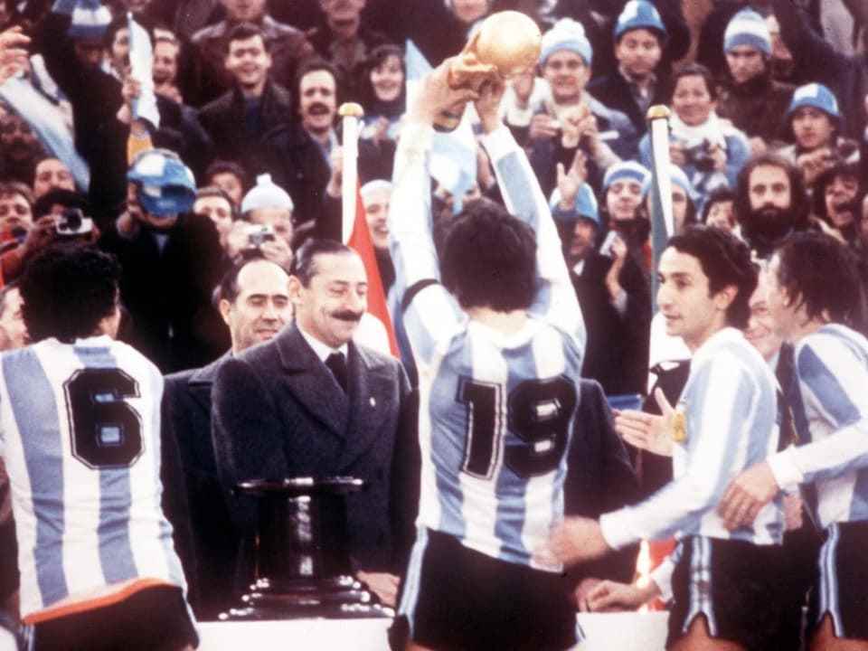 With their backs to the camera: four Argentinian players.  In the background Videla, who smiles a little with his coat and tie.
