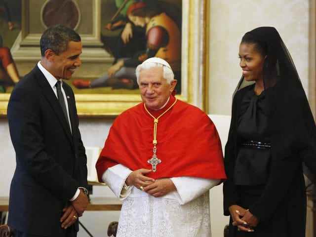 The Pope stands between the Obamas who wear black.  Michelle Obama wears a veil.