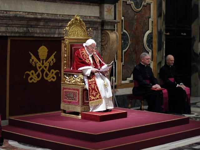 The Pope sits on a decorated chair and reads from a paper.