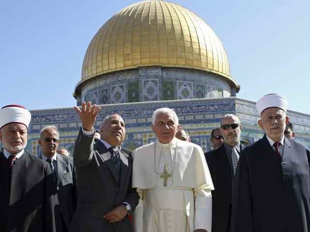 The Pope stands in front of the Dome of the Rock with other men in suits. 