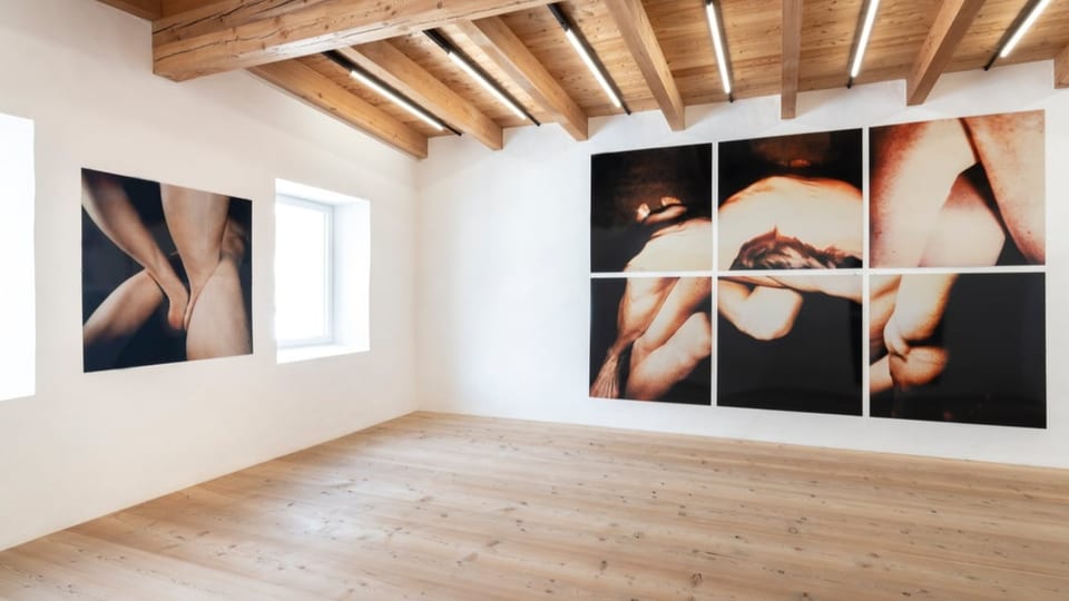 Room with white hand, wooden floor.  At the back on the wall is a large picture made up of six Polaroids, showing bodies in front of black paint