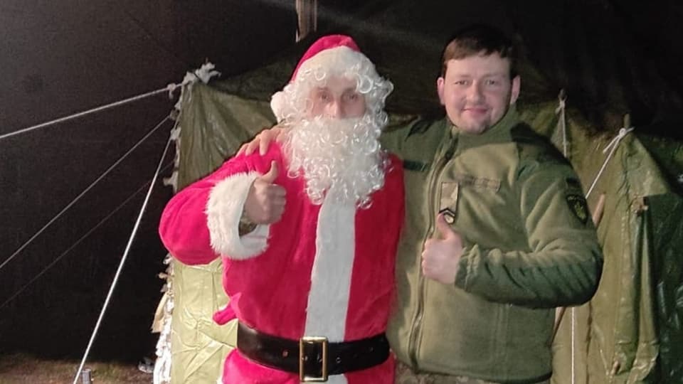 A soldier dressed as Santa Claus poses with a soldier in uniform in front of a military tent.
