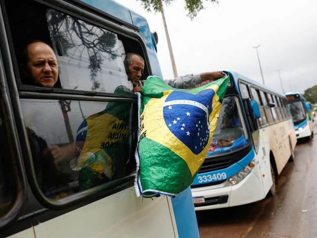 Men keep their heads out of a moving bus, one waving a Brazilian flag